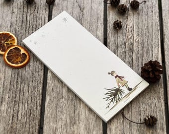 May include: A rectangular white notepad with a winter scene illustration. The illustration depicts a person carrying a Christmas tree. The notepad is decorated with small snowflakes. Dried orange slices and pinecones are scattered around the notepad.