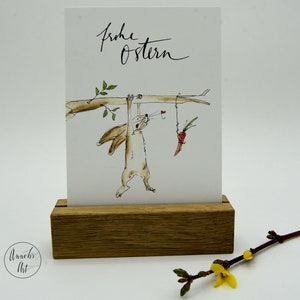 Puede incluir: Una ilustraci&oacute;n de acuarela de un conejo marr&oacute;n colgado boca abajo de una rama, alcanzando una zanahoria que cuelga de una cuerda. El texto "Frohe Ostern" est&aacute; escrito en caligraf&iacute;a negra en la parte superior de la tarjeta.