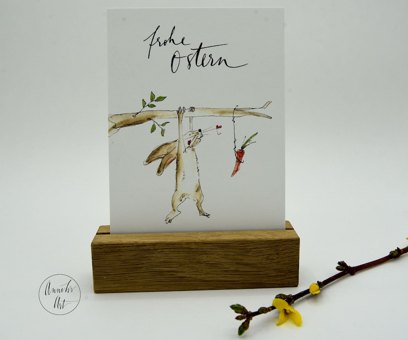 Puede incluir: Una ilustraci&oacute;n de acuarela de un conejo marr&oacute;n colgado boca abajo de una rama, alcanzando una zanahoria que cuelga de una cuerda. El texto "Frohe Ostern" est&aacute; escrito en caligraf&iacute;a negra en la parte superior de la tarjeta.
