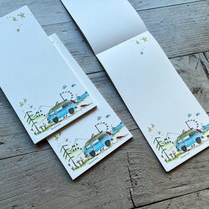 Könnte beinhalten: Drei weiße Papierblöcke mit einer farbenfrohen Illustration eines blauen Campingbusses in einer Landschaft mit grünen Bäumen, kleinen Pilzen und gelben Sternen. Das Design umfasst eine Wimpelkette über dem Bus sowie blaue Wolken.