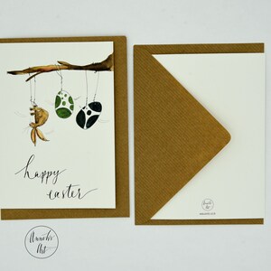 May include: A brown card with a white background and a watercolor illustration of a bunny hanging from a branch with two green and black Easter eggs. The text "happy easter" is written in black ink below the illustration.