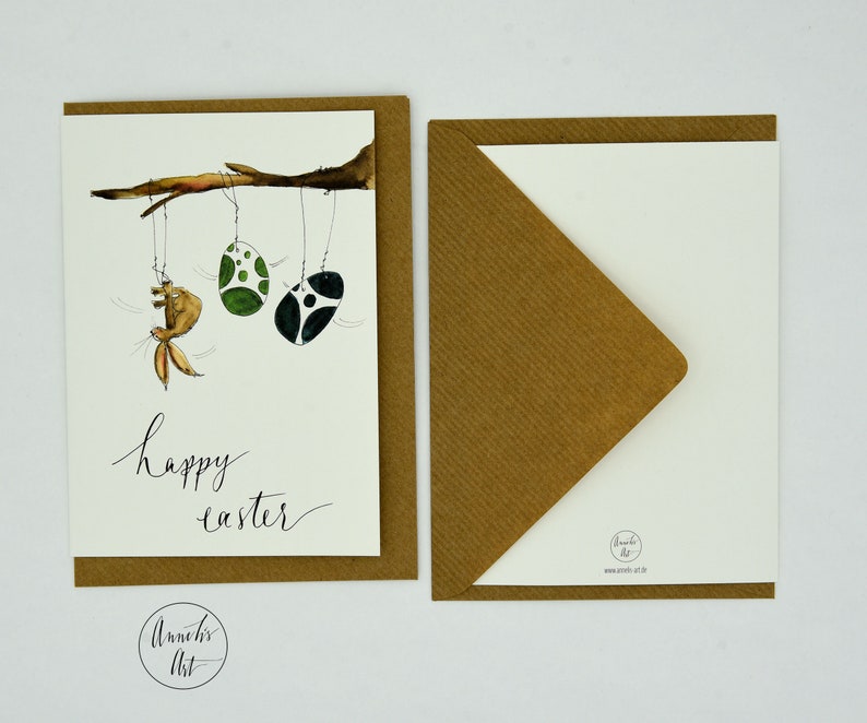 May include: A brown card with a white background and a watercolor illustration of a bunny hanging from a branch with two green and black Easter eggs. The text "happy easter" is written in black ink below the illustration.