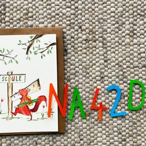 May include: A card with a cartoon squirrel carrying a ruler and a pencil, walking towards a sign that says "Schule" (German for school). The card is surrounded by colorful letters that spell out "NA42DE".