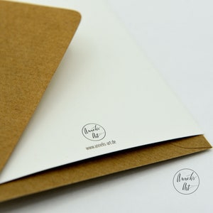 May include: A white card with a brown envelope. The card has a black circle with the text "Annelies Art" and the website address "www.annelies-art.de".