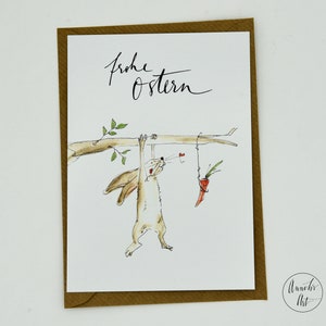 Puede incluir: Una tarjeta blanca con un borde marr&oacute;n presenta una ilustraci&oacute;n de acuarela de un conejito colgado de una rama y alcanzando una zanahoria. El texto "Frohe Ostern" est&aacute; escrito en caligraf&iacute;a negra en la parte superior de la tarjeta. La tarjeta est&aacute; firmada "Annelies Art" en la esquina inferior derecha.