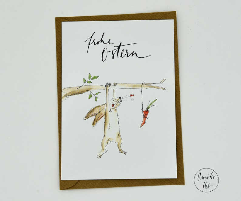Puede incluir: Una tarjeta blanca con un borde marr&oacute;n presenta una ilustraci&oacute;n de acuarela de un conejito colgado de una rama y alcanzando una zanahoria. El texto "Frohe Ostern" est&aacute; escrito en caligraf&iacute;a negra en la parte superior de la tarjeta. La tarjeta est&aacute; firmada "Annelies Art" en la esquina inferior derecha.