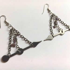May include: A pair of silver-toned earrings with a unique design. Each earring features a hook, a chain, and a series of decorative elements, including small charms and geometric shapes. The earrings have a modern, edgy aesthetic.