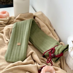 May include: A sage green hot water bottle with a ribbed texture, lying on a beige blanket. The bottle has a drawstring closure with a red cord and a wooden bead. A small wooden tag is attached to the bottle.