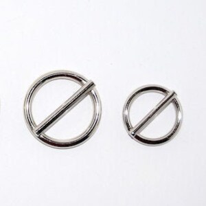 Puede incluir: Cuatro anillos met&aacute;licos plateados con una barra diagonal en el centro. Los anillos son de diferentes tama&ntilde;os, dispuestos de mayor a menor, sobre un fondo blanco. Los anillos parecen estar hechos de un metal brillante y reflectante.