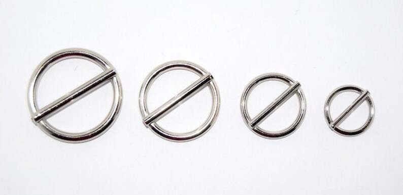 Puede incluir: Cuatro anillos met&aacute;licos plateados con una barra diagonal en el centro. Los anillos son de diferentes tama&ntilde;os, dispuestos de mayor a menor, sobre un fondo blanco. Los anillos parecen estar hechos de un metal brillante y reflectante.