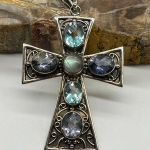 May include: A silver cross pendant with five gemstones. The center stone is a large, iridescent labradorite. The other four stones are smaller and are a mix of blue topaz and amethyst.