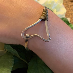 May include: A silver heart-shaped bangle bracelet with a gold wire coil accent. The bracelet is worn on a person's wrist.