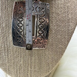 May include: A silver-colored pendant necklace with a rectangular pendant featuring an engraved floral and starburst design. The pendant is approximately 2 inches long. The necklace has turquoise-colored beads.