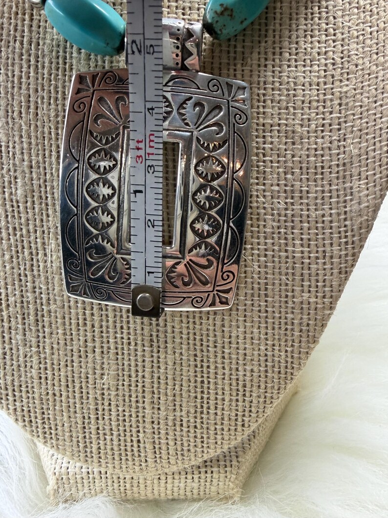 May include: A silver-colored pendant necklace with a rectangular pendant featuring an engraved floral and starburst design. The pendant is approximately 2 inches long. The necklace has turquoise-colored beads.