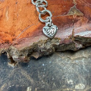May include: Silver-toned bracelet with a heart-shaped charm. The charm features a cursive "B" inside the heart. The bracelet is composed of interconnected circular links. The background is a mix of brown and gray tones.