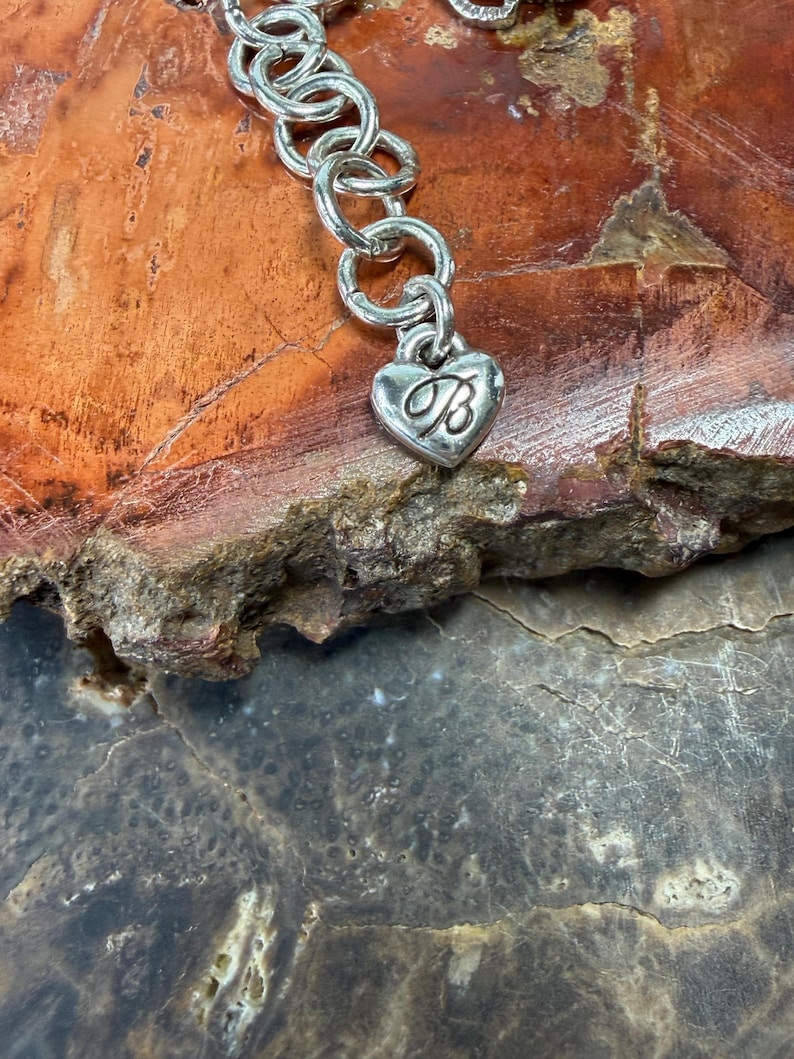 May include: Silver-toned bracelet with a heart-shaped charm. The charm features a cursive "B" inside the heart. The bracelet is composed of interconnected circular links. The background is a mix of brown and gray tones.