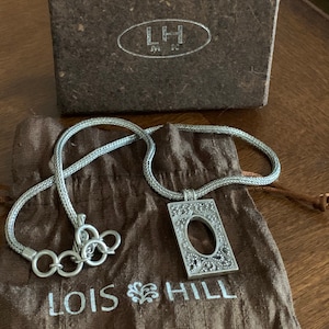 May include: A silver chain necklace with a silver pendant featuring an ornate design. The pendant is rectangular with a cutout oval in the center. The necklace is displayed on a brown fabric pouch with the text "Lois Hill" printed on it.
