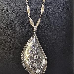 May include: A silver necklace with a diamond-shaped pendant featuring a detailed leaf design and three clear stones.