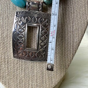 May include: Silver-tone rectangular pendant necklace with turquoise and copper-colored beads. The pendant is approximately 2 inches long and features an ornate, engraved design. The necklace is displayed on a beige fabric surface.