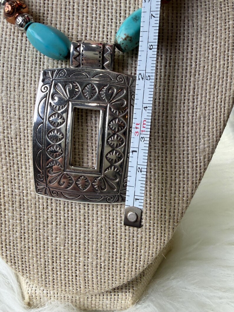 May include: Silver-tone rectangular pendant necklace with turquoise and copper-colored beads. The pendant is approximately 2 inches long and features an ornate, engraved design. The necklace is displayed on a beige fabric surface.