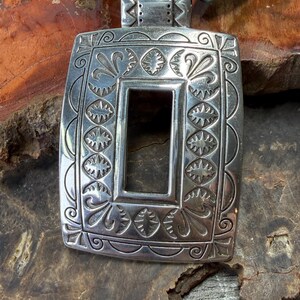 May include: A silver-toned rectangular pendant with intricate embossed floral and geometric patterns. The pendant has a central rectangular cutout and is strung on a turquoise beaded necklace. The pendant is displayed on a textured, brown surface.