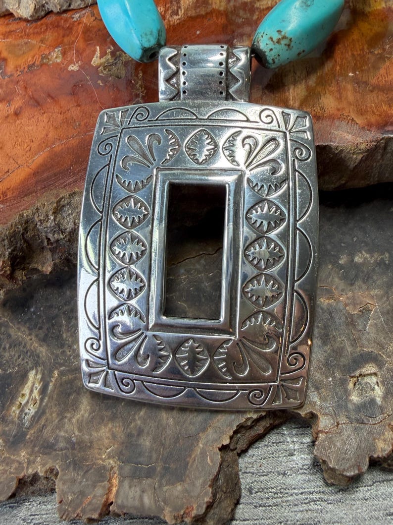 May include: A silver-toned rectangular pendant with intricate embossed floral and geometric patterns. The pendant has a central rectangular cutout and is strung on a turquoise beaded necklace. The pendant is displayed on a textured, brown surface.