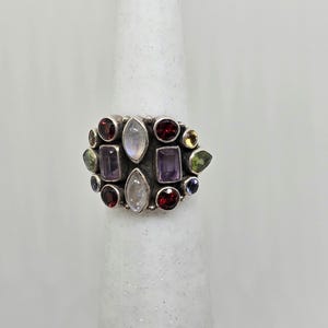 May include: A silver ring featuring an array of colorful gemstones. The ring showcases a cluster of various shapes and sizes of stones, including red, purple, green, and iridescent white gems. The ring is set on a white background.