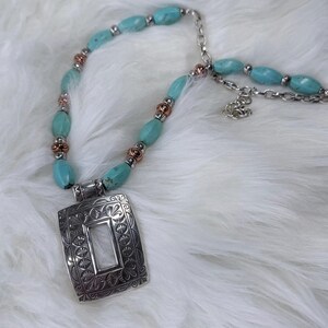 May include: A turquoise beaded necklace with a silver pendant. The pendant is rectangular with an ornate floral design and a white center. The necklace features turquoise oval beads, silver chain, and copper-colored accents.