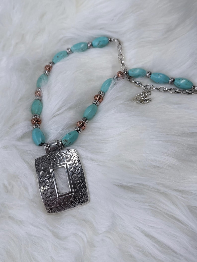 May include: A turquoise beaded necklace with a silver pendant. The pendant is rectangular with an ornate floral design and a white center. The necklace features turquoise oval beads, silver chain, and copper-colored accents.