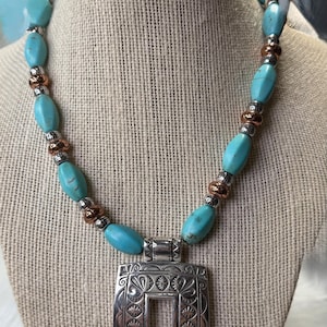 May include: A turquoise beaded necklace with a silver pendant. The necklace features oval turquoise beads, copper-colored spacers, and a rectangular silver pendant with an ornate floral design. The pendant has a rectangular cutout in the center.