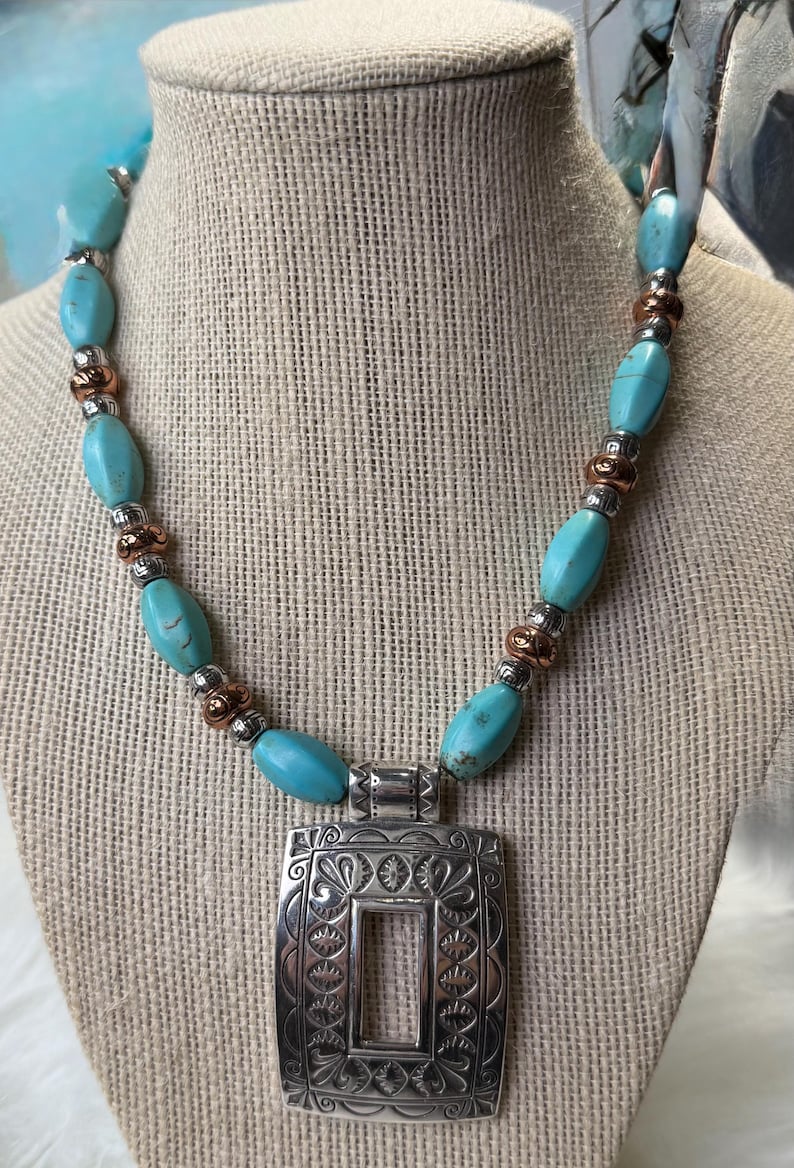 May include: A turquoise beaded necklace with a silver pendant. The necklace features oval turquoise beads, copper-colored spacers, and a rectangular silver pendant with an ornate floral design. The pendant has a rectangular cutout in the center.