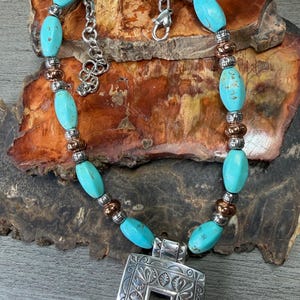 May include: Turquoise beaded necklace with a silver pendant. The necklace features oval turquoise beads, silver and copper-colored accents, and a rectangular pendant with an ornate design. The necklace is displayed on a wooden surface.