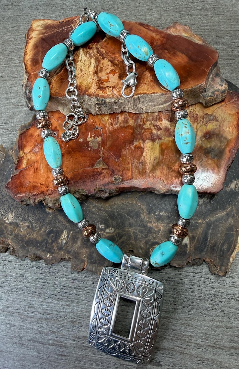 May include: Turquoise beaded necklace with a silver pendant. The necklace features oval turquoise beads, silver and copper-colored accents, and a rectangular pendant with an ornate design. The necklace is displayed on a wooden surface.