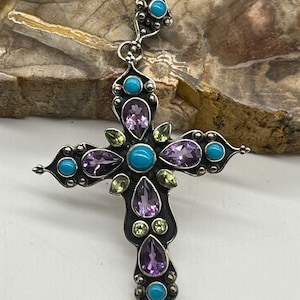 May include: A silver cross pendant with turquoise, amethyst, and green gemstones. The cross is ornate and detailed, with a bail at the top for hanging.