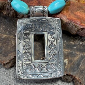 May include: Silver-tone pendant necklace with turquoise and silver beads. The rectangular pendant has an open center and is decorated with intricate patterns. The necklace is displayed on a textured, brown surface.