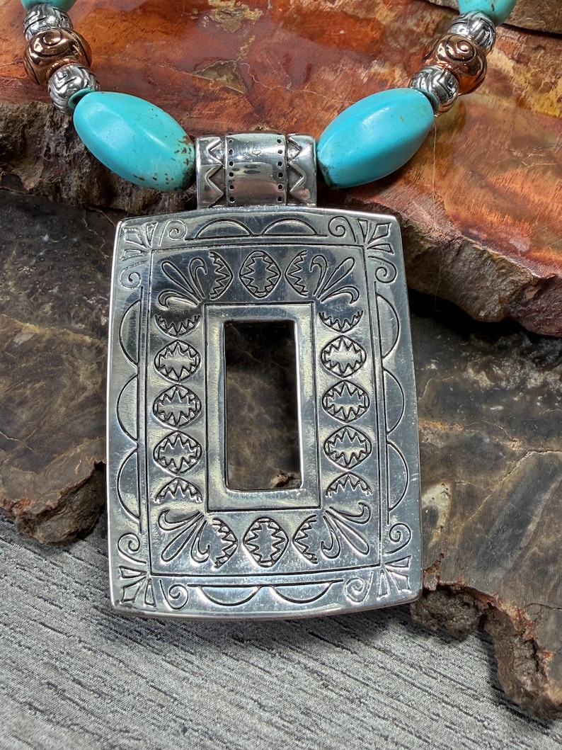 May include: Silver-tone pendant necklace with turquoise and silver beads. The rectangular pendant has an open center and is decorated with intricate patterns. The necklace is displayed on a textured, brown surface.