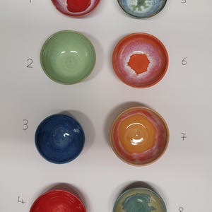 Könnte beinhalten: Acht Keramikschalen in zwei Spalten angeordnet, nummeriert von 1 bis 8. Die Schalen weisen verschiedene Glasurfarben auf, darunter Rot, Grün, Blau, Orange und mehrfarbige Mischungen mit weißen Zentren. Jede Schale hat eine glatte Innenfläche.