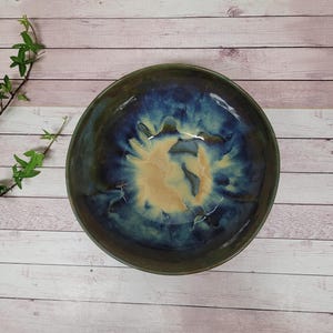 Könnte beinhalten: Eine handgefertigte Keramikschale mit einer einzigartigen Glasur. Die Schale weist eine Mischung aus tiefblauen, grünen und beigen Farben auf, wodurch ein marmorierter Effekt entsteht. Das Innere hat eine hellere, beige Mitte, während der Rand ein dunkleres Blaugrün ist. Die Schale steht vor einem hellen Holzhintergrund.