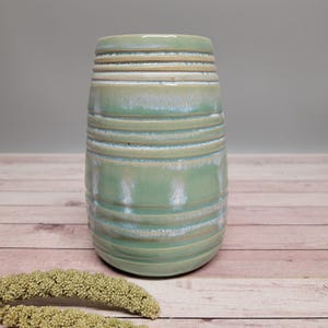 Könnte beinhalten: Eine zylindrische, handgefertigte Keramikvase mit hellgrüner Glasur. Die Vase hat horizontale, strukturierte Bänder um ihren Körper. Die Vase steht auf einer Holzoberfläche mit getrockneten Pflanzenstielen.