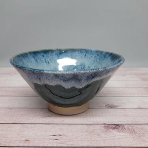 Könnte beinhalten: Handgefertigte Keramikschale mit blau-weißer Glasur. Die Schale hat eine runde Form mit leicht ausgestelltem Rand und beigem Sockel. Das Innere der Schale ist dunkler blau.