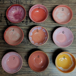 Könnte beinhalten: Neun Keramikschalen in verschiedenen Farben, angeordnet auf einer Holzoberfläche. Die Schalen sind von 1 bis 9 nummeriert. Die Farben umfassen Rot-, Rosa-, Orange- und Gelbtöne. Jede Schale hat ein einzigartiges Design und eine Farbvariation.