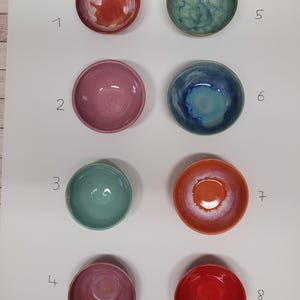 Könnte beinhalten: Acht kleine Keramikschalen in zwei Spalten angeordnet, nummeriert von eins bis acht. Die Schalen haben verschiedene glänzende Glasuroberflächen in Rot-, Rosa-, Türkis-, Blau- und Orangetönen, jede mit einzigartigen Farbverläufen und Texturen.