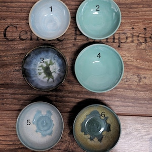 Könnte beinhalten: Sechs kleine, handgefertigte Keramikschalen in verschiedenen Blau-, Grün- und Brauntönen. Jede Schale hat ein einzigartiges Glasurmuster, einige mit Wirbel- oder Sprenkeldesigns. Die Schalen sind auf einer Holzoberfläche angeordnet und zeigen ihre individuellen Farben und Texturen.