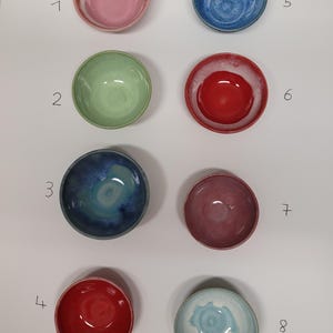 Könnte beinhalten: Acht kleine Keramikschalen in zwei Spalten angeordnet, nummeriert von eins bis acht. Die Schalen weisen verschiedene Glasurfarben auf, darunter Rosa, Hellgrün, Dunkelblau, Rot, Hellblau und Mauve, jeweils mit einzigartigen Farbnuancen.