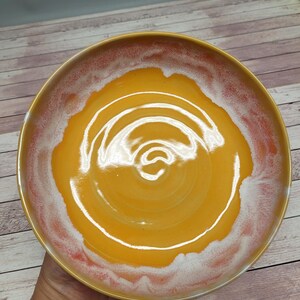 Könnte beinhalten: Eine handgefertigte Keramikschale mit einem leuchtend gelben Zentrum und einem Farbverlauf am Rand. Der Rand geht von rot-orange nach weiß über und erzeugt einen einzigartigen visuellen Effekt. Das Design der Schale ist schlicht und elegant, geeignet zum Servieren oder Ausstellen.