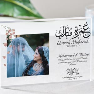 May include: A clear acrylic photo frame with a floral design and a picture of a couple. The frame has Arabic calligraphy that reads "Umrah Mubarak" and "December 2024". The text below the calligraphy reads "Mohammed & Fatima, May Allah accept your Umrah, and shower you with His blessings. Ameen. "And we have created you in pairs" Quran 78:8".