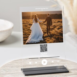 Puede incluir: Un marco de fotos de acrílico transparente con una base negra. El marco muestra una foto de una pareja caminando hacia atrás en un campo. El marco también incluye un código QR, el texto "Perfect" y "Ed Sheeran", y una interfaz de reproductor de música con botones de reproducción, pausa y avance.