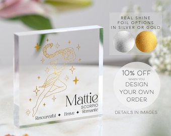 May include: A clear acrylic block with a gold foil design of a woman with a star-shaped halo. The text "Mattie Scorpio Resourceful Brave Romantic" is also in gold foil. The block is on a white surface.