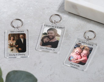 May include: Three clear acrylic keychains with photos and personalized messages. The first keychain has a photo of a father and daughter walking down the aisle, with the text "Father & Daughter" and "Emily & Daddy". The second keychain has a photo of a father and daughter hugging, with the text "To the best dad in the world" and "Love Amelia xx". The third keychain has a photo of two young girls hugging, with the text "This daddy is loved by" and "Ava & Ella".
