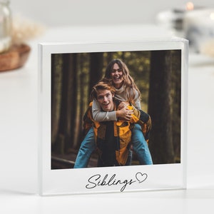 Puede incluir: Un cubo de foto de acrílico blanco con una imagen de dos hermanos en un bosque. La niña está en la espalda del niño y ambos están sonriendo. El texto "Siblings ♥" está impreso en la parte inferior del cubo.